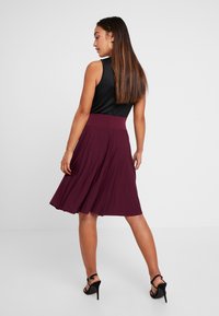 Saia plissada cor burdeos com um cós largo, combinada com uma blusa preta sem mangas e sandálias pretas de salto alto. Textura de tecido suave.