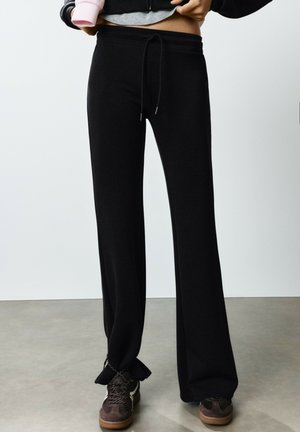 Personne portant un pantalon noir à jambes larges avec une taille à cordon et des baskets marron, debout sur un sol gris contre un mur blanc.
