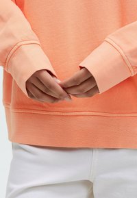Bright orange sweatshirt with a relaxed fit and ribbed cuffs, paired with white pants. The fabric has a soft, textured appearance.