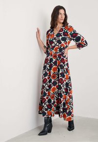 Žena u dugoj cvjetnoj haljini sa crvenim, narančastim, ljubičastim i zelenim cvjetovima, pozira s jednom rukom na zidu i crnim čizmama.