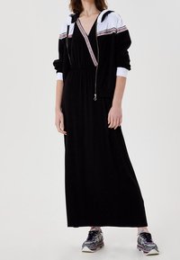 Schwarzes Maxikleid mit geraffter Taille, kombiniert mit einer schwarz-weißen Zip-up-Kapuzenjacke mit dekorativem Band. Gekleidet mit gemusterten Sportsneakern.