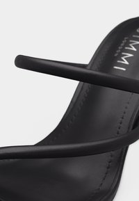 Μαύρο σανδάλι με ανοιχτό δάχτυλο και δύο λεπτές λουρίδες που διασχίζουν το πόδι, με επίπεδη σόλα και μερική εμπορική σήμανση στην εσωτερική σόλα.