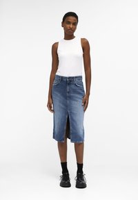 Haut sans manches blanc et jupe en denim mi-longue avec une fente à l'avant. Des chaussures noires et des chaussettes à mi-mollet complètent la tenue.