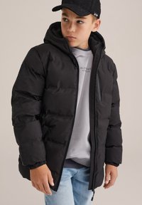 Juodas pūkinis džemperis su gobtuvu, su užtrauktuku priekyje ir elastinėmis rankovėmis. Dėvimas ant pilkos sportinės megztinės ir džinsų.