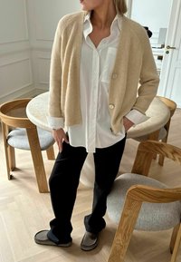 Bézs gyapjú kardigán kör alakú gombokkal, fehér inggel, fekete nadrággal és szürke papuccsal. Puha tapintású és laza fazonú. Egyszerű háttér.