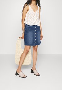 Žena v bílém vzorovaném bezramínkovém topu, denimové sukni s knoflíky, béžových vysokých mule, držící velkou pletenou tašku.