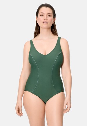 Mujer con el cabello mojado que lleva un traje de baño entero verde oscuro con tirantes anchos, posando frente a un fondo claro y sencillo.