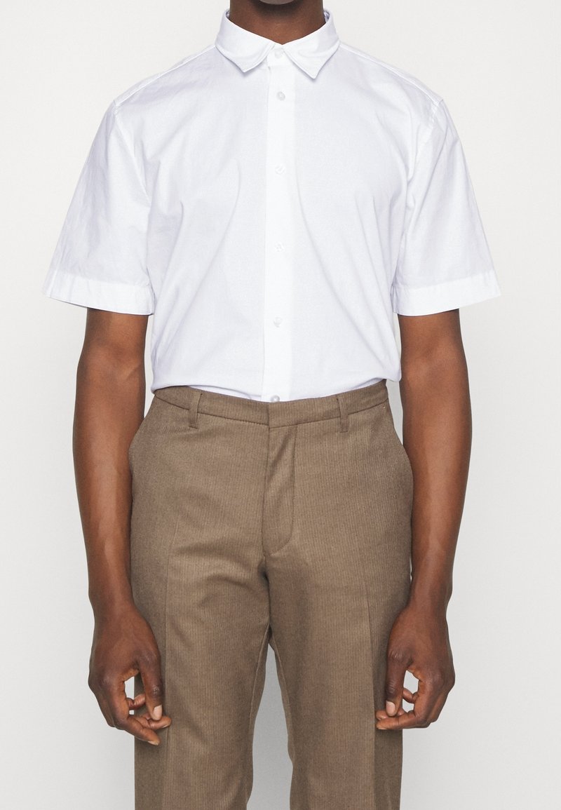 Camisa de algodón blanca de manga corta con cuello, con botones en la parte delantera, llevada con pantalones marrones texturizados.