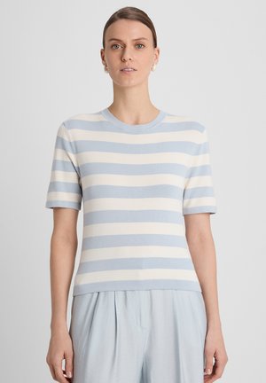 Femme portant un t-shirt à manches courtes rayé horizontalement bleu clair et blanc, et un pantalon bleu clair, debout devant un fond clair uni.
