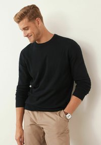 Schwarzer Rundhals-Pullover aus weichem Material, kombiniert mit beigen Hosen. Auffällige Details sind gerippte Bündchen und Saum. Silberne Uhr sichtbar.