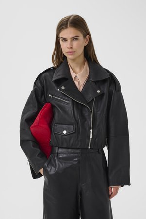 Noor naine kannab musta nahktagi ja pükse, hoides punast tekstuuriga käekotti heleda ühevärvilise tausta ees.