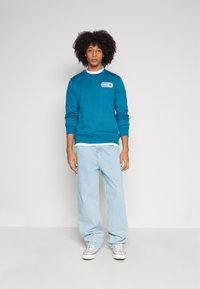 Türkisfarbener Sweatshirt mit Rundhalsausschnitt und einem Patches auf der Brust, kombiniert mit hellblauen, lässig geschnittenen Jeans und weißen Sneakern.