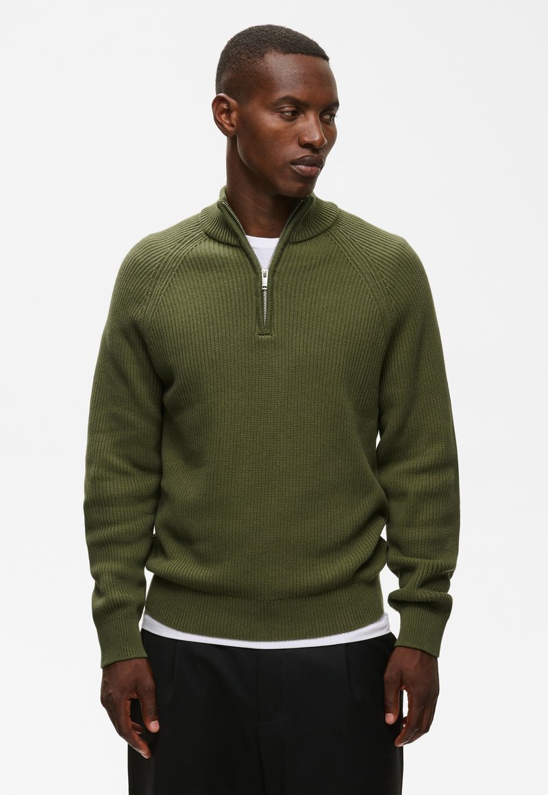 Homme portant un pull en maille côtelée vert olive avec un col demi-zip sur une chemise blanche, debout devant un fond blanc uni.