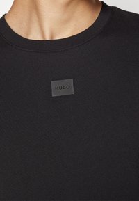 Vicinanza di una persona che indossa una maglietta nera a girocollo con un piccolo patch rettangolare al centro del petto che recita "HUGO".