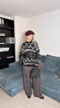Maglione a righe nere e grigie, pantaloni grigi a gamba larga, berretto bordeaux, mentre tiene una borsa nera. In piedi su un divano blu con texture.