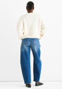 Cremefarbener Pullover mit strukturierten Noppen, lockere Passform. Blaue hoch geschnittene weit geschnittene Jeans mit Verwaschungen und Gesäßtaschen. Schwarze flache Schuhe.