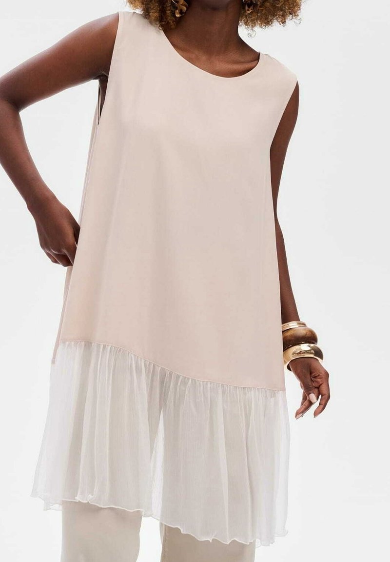 Femme portant une robe beige sans manches avec un ourlet à volants blanc transparent et de gros bracelets dorés au poignet droit, sur un fond uni.
