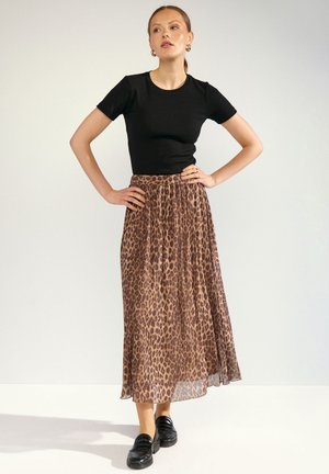 Donna in piedi con le mani sui fianchi, indossa una maglia nera a maniche corte, una gonna lunga con stampa leopardata e scarpe nere slip-on.