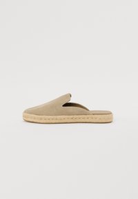 Bežs zamša slip-on kurpes ar atvērtu aizmuguri un sānu izgriezumiem, ar dabīgu jutu virsu un plakanu dizainu.