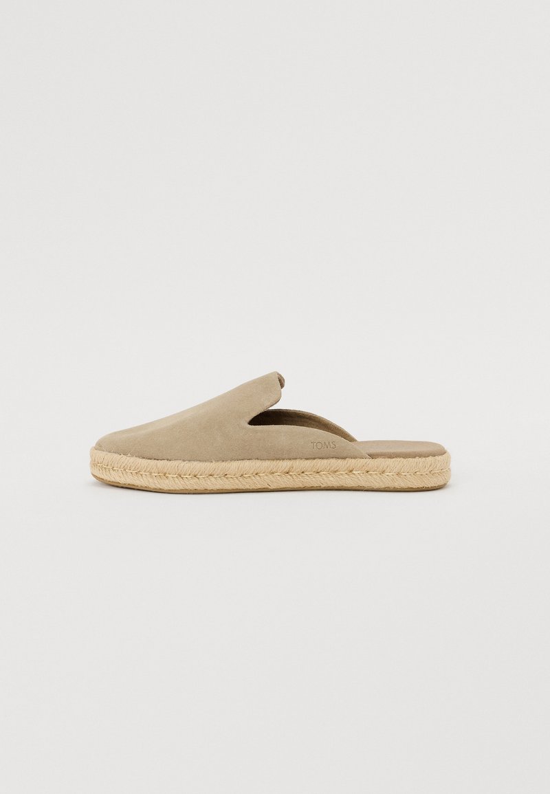 Bežs zamša slip-on kurpes ar atvērtu aizmuguri un sānu izgriezumiem, ar dabīgu jutu virsu un plakanu dizainu.