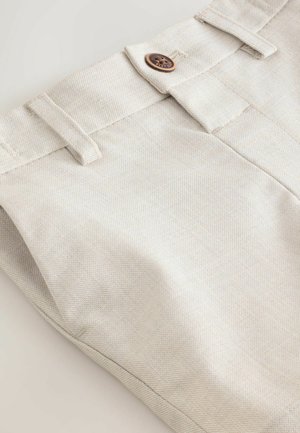 Beige katoenen broek met een gestructureerde afwerking, voorzien van een bruine knoop en twee riemlussen in de tailleband. Detail van een onafgewerkte zoom zichtbaar.