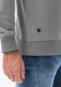 Uomo in maglione grigio a maniche lunghe con toppa logo e braccialetto di perline blu, jeans di denim blu, mano rilassata lungo il fianco.
