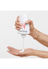 Kérastase haarproduct in een witte fles met roze accenten die een witte romige textuur in een open hand giet, met een strak ontwerp.