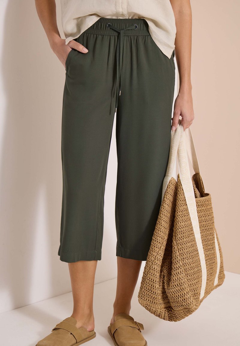 Femme portant un pantalon court vert foncé avec une taille à cordon, un haut beige, des sandales beiges à enfiler et tenant un grand sac fourre-tout tissé.