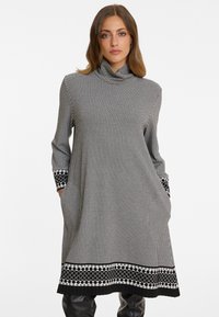 Schwarz-weiß kariertes Kleid mit hohem Kragen, langen Ärmeln, seitlichen Taschen und einem gemusterten Saum mit kreisförmigen Akzenten.