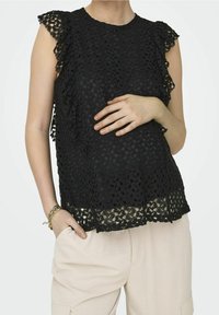 Top in pizzo nero con un design testurizzato, maniche a volant e orlo scallop, indossato sopra pantaloni beige chiaro.