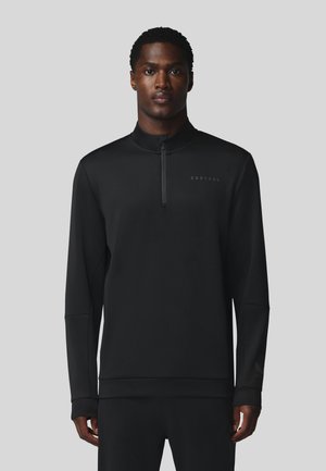 Man die naar voren kijkt, draagt een zwarte lange mouwen quarter-zip trui met het "Castore" logo op de borst en zwarte broek.