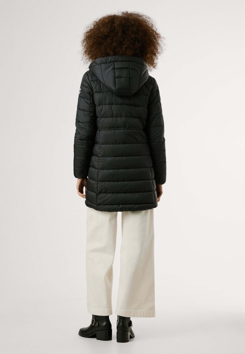 Cappotto nero imbottito con cappuccio, caratterizzato da un design trapuntato e tasche laterali, abbinato a pantaloni bianchi a gamba larga e stivaletti neri con tacco.