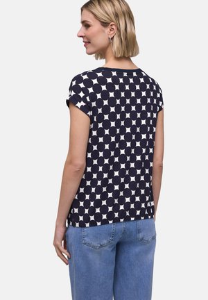 Vrouw draagt een navyblauw-wit polkadot-shirt met korte mouwen en omgeslagen mouwen en lichtblauwe spijkerbroek, gezien van achteren, licht glimlachend.
