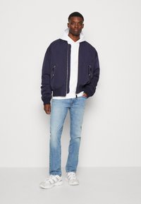 Marine bomberjakke med glidelåslommer, båret over en hvit hettegenser. Lyseblå denimjeans og grå sneakers fullfører antrekket.