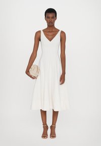 Witte, mouwloze A-lijn jurk met een V-hals, voorzien van verticale naden en een vloeiende zoom. Model houdt een kleine beige tas vast. Bruine hakken maken de look compleet.