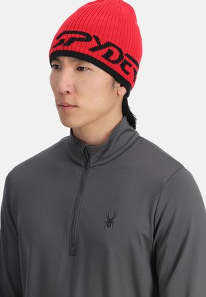 Rode gebreide muts met zwart merk, gedragen met een grijs, lang mouw zip-up shirt met een klein spinnenlogo op de borst.