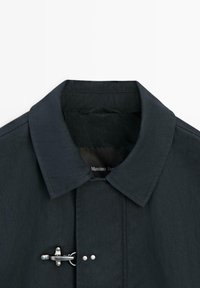 Colletto di cappotto blu navy scuro con chiusura a tirante metallico e due bottoni automatici, con cuciture visibili e etichetta del marchio parzialmente visibile all'interno.