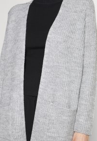 Jasnoszary dzianinowy kardigan z otwartym przodem i kieszeniami, noszony na czarnym prążkowanym topie z długim rękawem i czarnych spodniach.