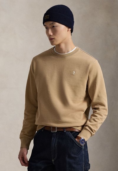 Felpe color camoscio con maniche lunghe, collo a costine e piccolo logo. Abbinate a jeans scuri e un berretto a maglia blu navy. Cintura in pelle marrone.