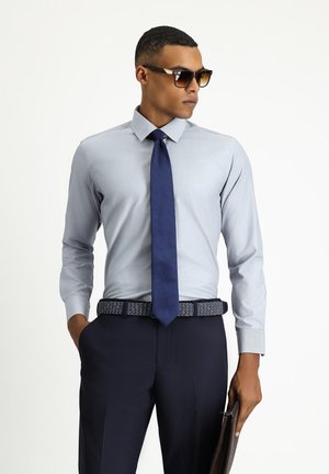 Camicia azzurro chiaro con colletto a punta, cravatta blu navy, cintura scura con motivo e pantaloni scuri. Gli accessori includono occhiali da sole e una cartella marrone.