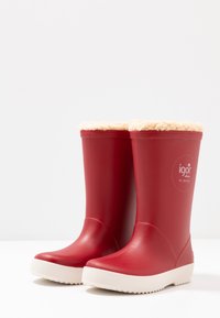 Bottes de pluie en caoutchouc rouges avec des semelles texturées blanches et un intérieur moelleux beige en haut. Logo de la marque visible sur le côté extérieur.