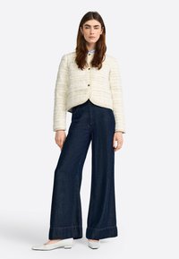 Giacca bianca in tweed con un taglio corto e chiusura a bottoni, abbinata a jeans in denim scuro a gamba larga e scarpe bianche. Texture liscia, pattern a quadretti.
