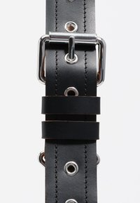 Črn usnjen trak s srebrno strojno opremo, ki vsebuje perforirane luknje in dva horizontalna usnjena traka za prilagajanje. Vidna podrobnost šivanja.