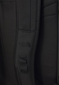 Cinghia di zaino nera realizzata in nylon testurizzato, dotata di una toppa con logo e di un anello in tessuto per il fissaggio. Design semplice e funzionale.