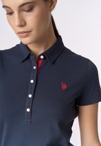 Polo shirt blu navy con un accento rosso sulla patta. Presenta un colletto, chiusura con bottoni e logo ricamato sul petto. Materiale in cotone.