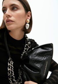 Eine Frau in einem schwarzen Oberteil hält eine strukturierte schwarze Tasche mit einer silbernen Kette. Sie trägt große goldene und silberne Ohrringe und sieht nachdenklich aus.