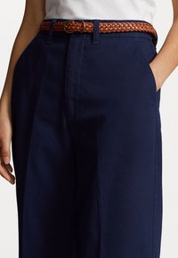 Mörkblå byxor med slät front, sidofickor och ett flätat brunt läderbälte med en rund metallspänne.