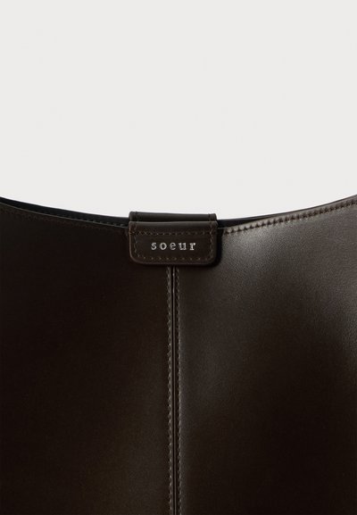 Gros plan d'un sac en cuir marron foncé avec une couture verticale et une petite étiquette cousue embossée avec "soeur" en haut au centre.