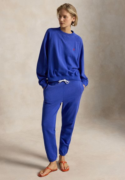 Kobieta stoi z rękami w kieszeniach, ubrana w niebieską bluzę i spodnie dresowe z białym sznurkiem oraz pomarańczowymi sandałami na neutralnym tle.