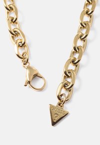 Colar de corrente dourada com elos oversized, fecho de lagosta e um charme triangular gravado com "GUESS". Textura suave e brilhante.